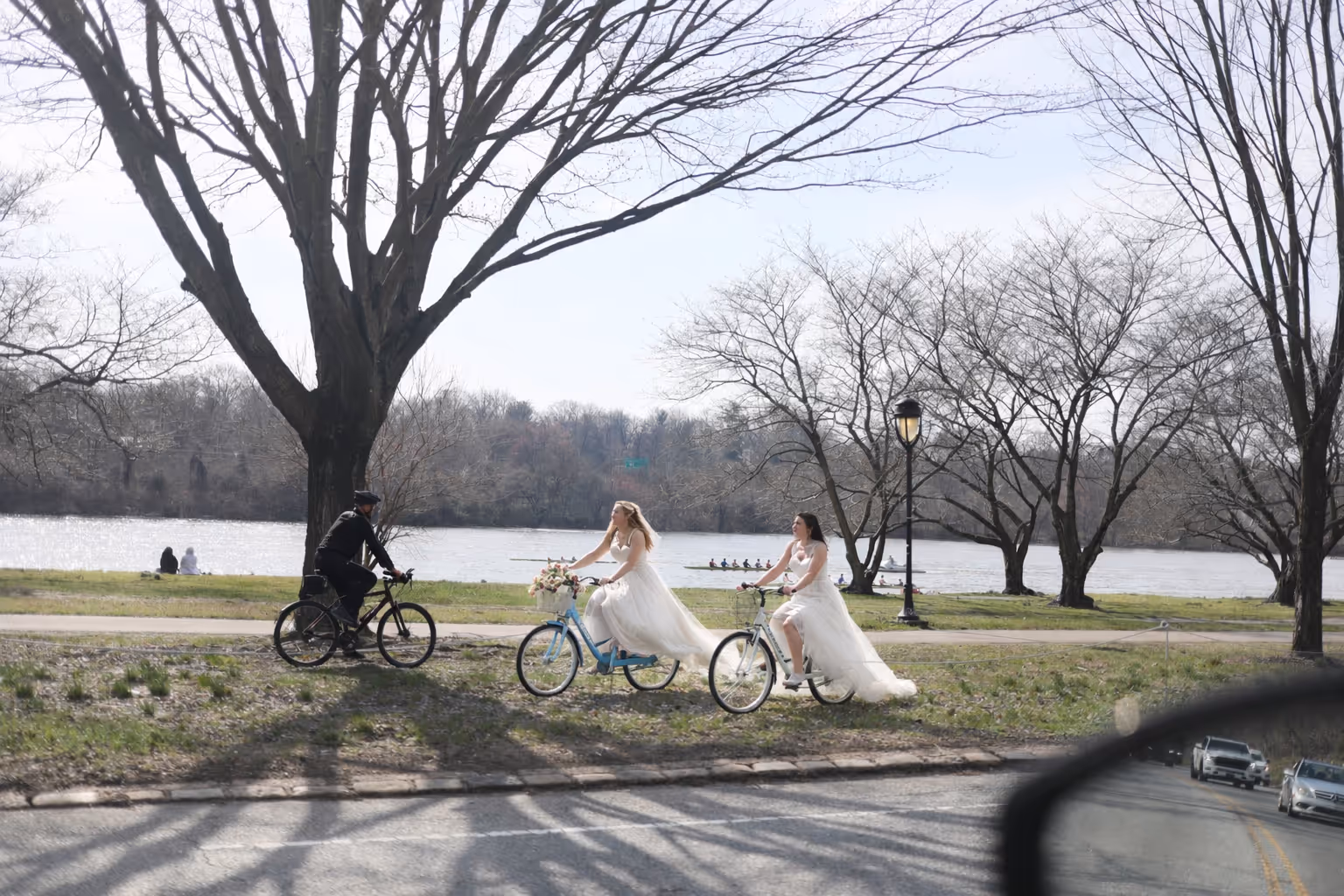 Two brides in wedding dresses riding bicycles near a river with leafless trees and a man on a bike approaching them.