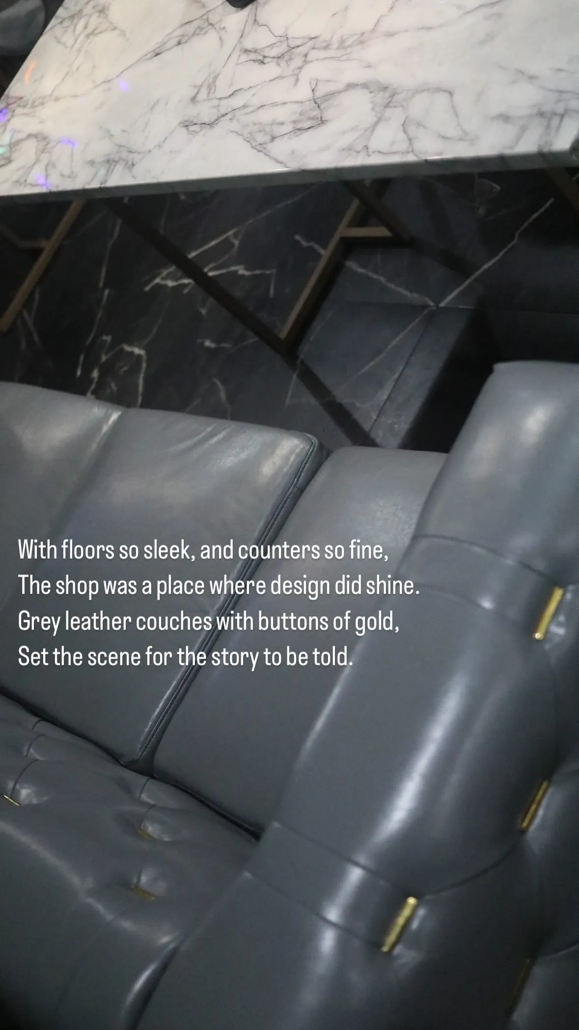 Close-up of grey leather couch with gold button accents beneath a white marble table on a dark marbled floor.